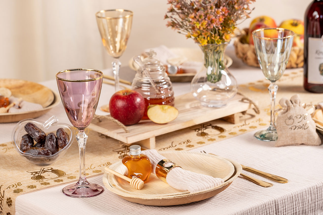 Table set for a meal with purple glassware, honey, dates, apple slices, and flowers on a patterned tablecloth